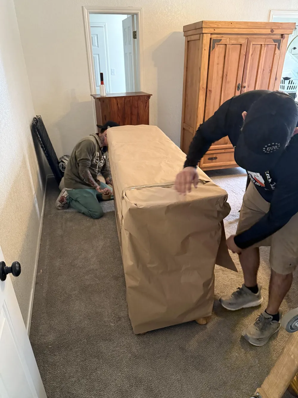 Two of the crew wrap a dresser in brown paper pads in a bedroom